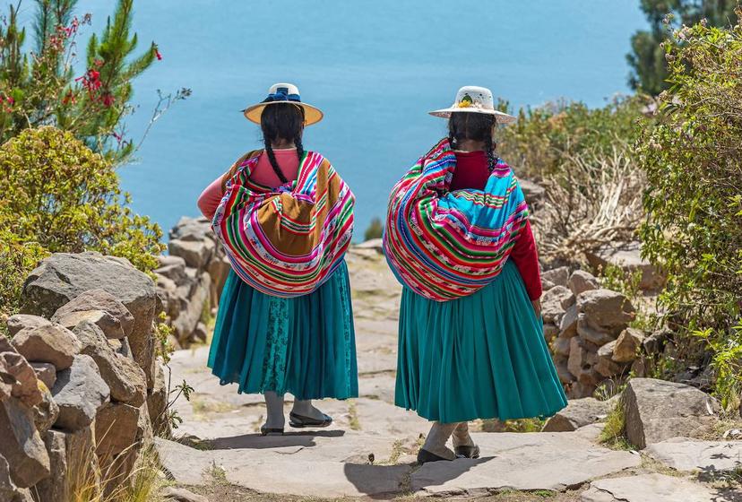 Two Quechua women walk down a path on Taquile Island with Lake Titicaca in the background. Photo by <a class="text-secondary" href="https://www.peruforless.com/">Peru For Less</a>