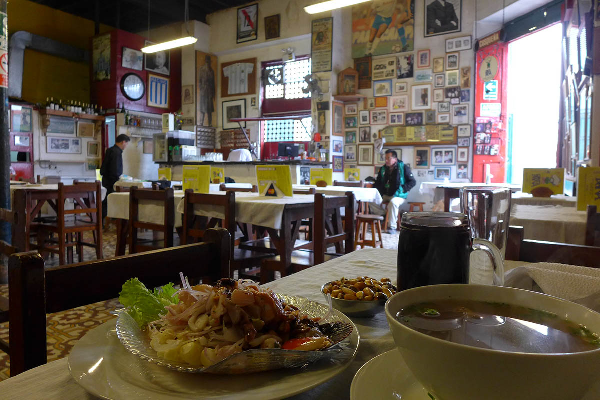 A soup bowl and plate of ceviche on a white clothed table. A wall covered by framed photos behind.