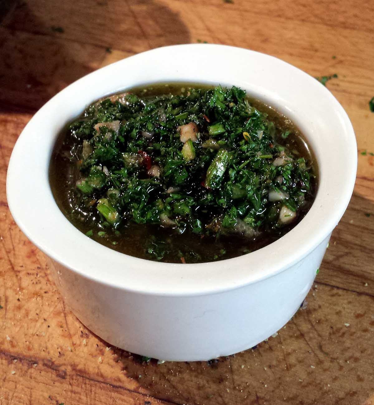 Green chimichurri sauce in a white bowl on a wood table.