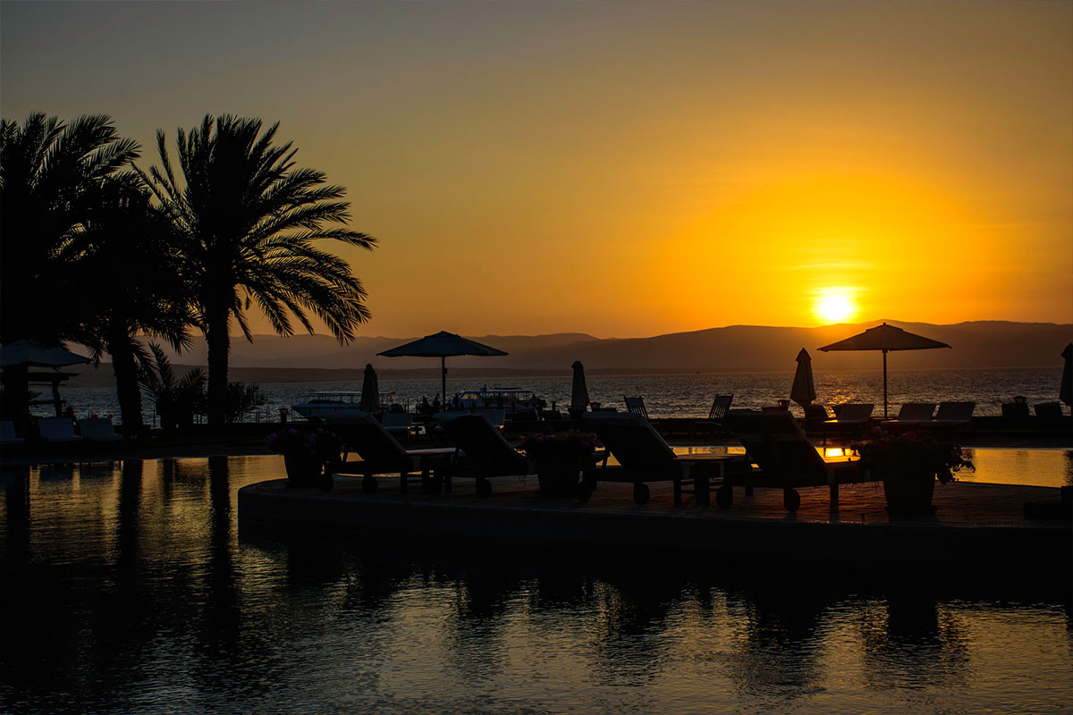 An orange and yellow sunset over the Paracas Peninsula with umbrella and palm tree silhouettes.