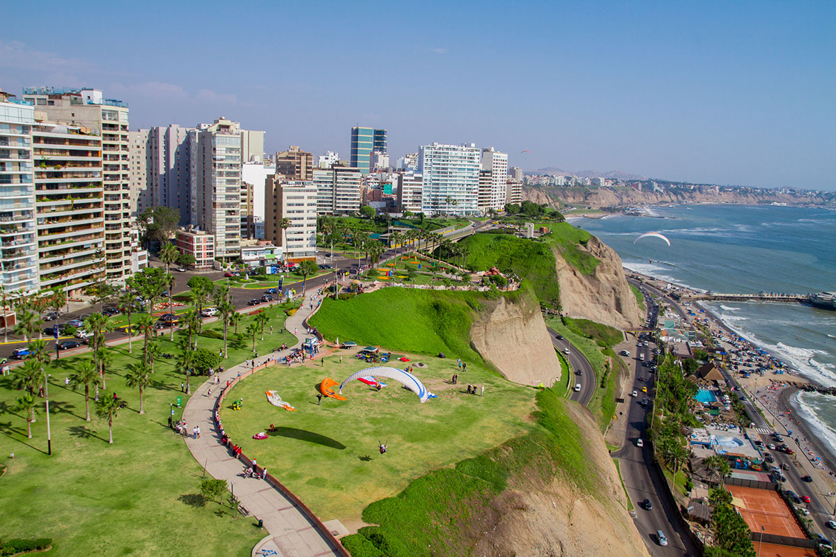 A low aerial view of the Malecon in Lima, Peru. There are many people walking on the Malecon, and along is green space and paragliders