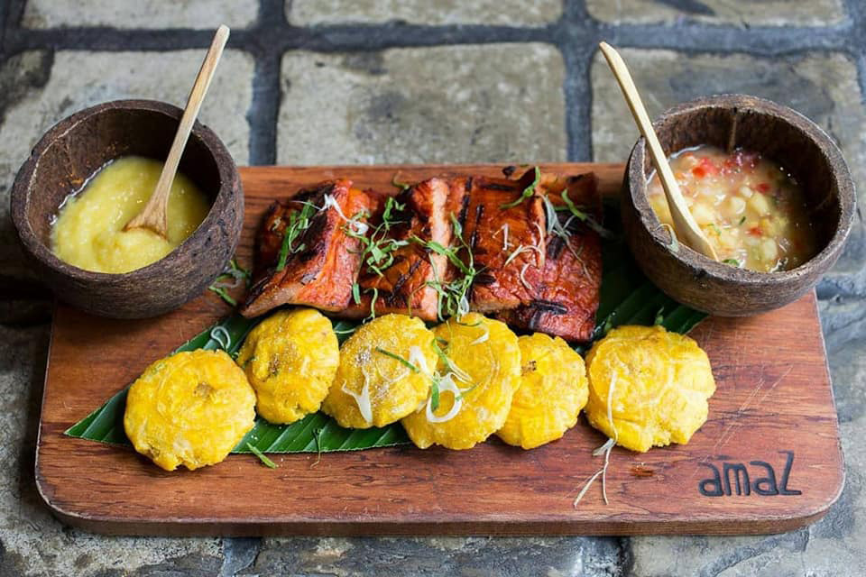 A wooden plate with a line of mashed plantains and pork served with two spicy sauces.