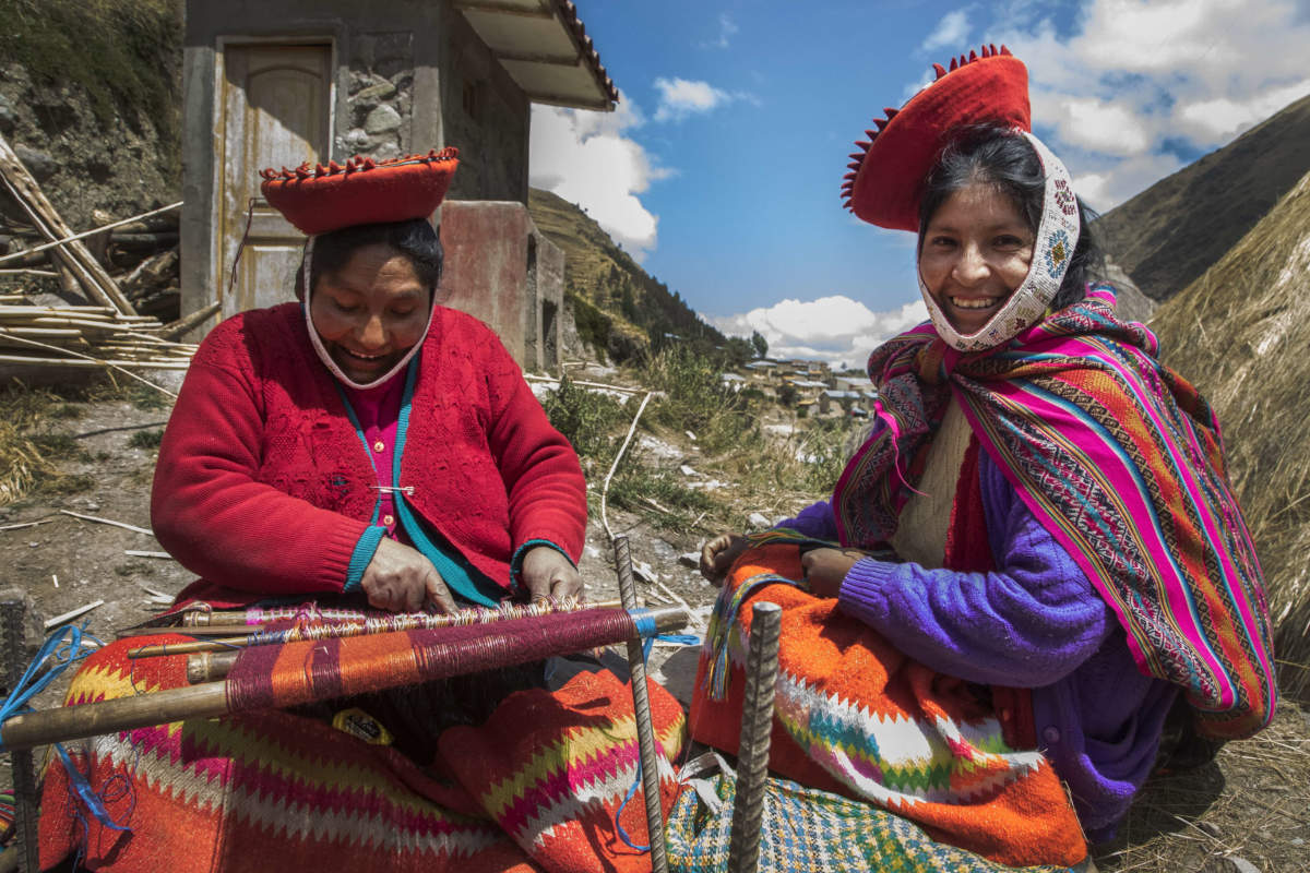 Two Andean women in traditional attire weaving textiles outside in the Peruvian Andes.