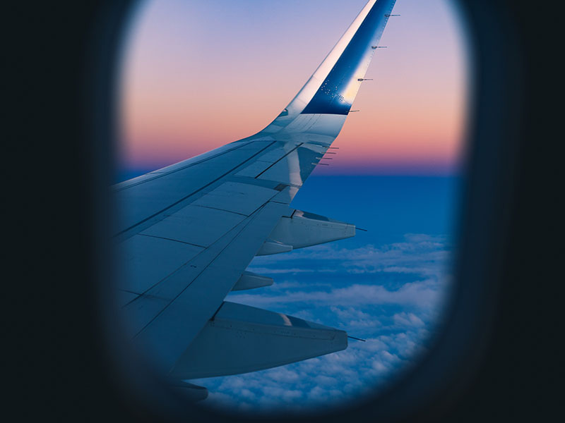 A window looking out at the wing of a plane as it flies over some clouds during sunset.