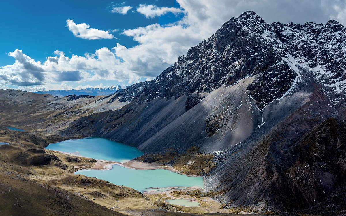 The Andes Mountains on a partly cloudy day with turquoise lakes at the base of a grey peak.