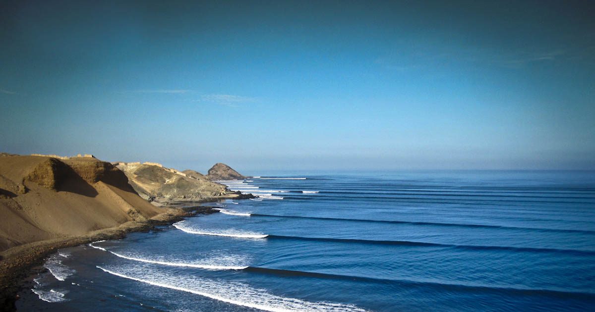 A wave breaks far in the distance with sandy cliffs and a blue sky in the background.