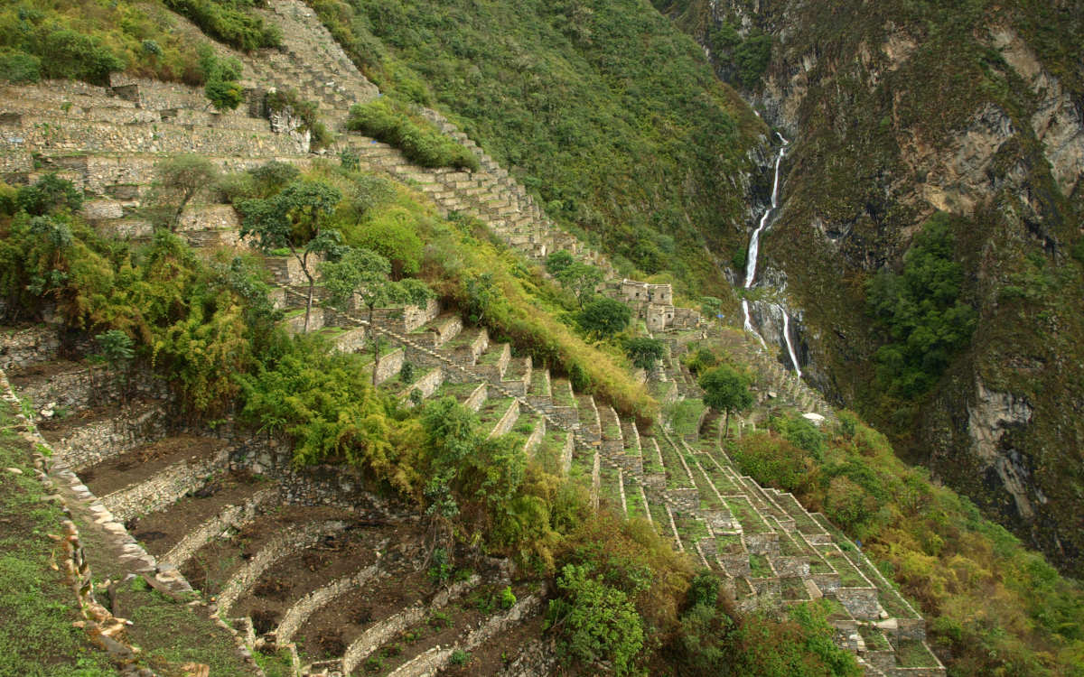 The lush ruins of Choquequirao nestled on the green Andean hillside with a cascade in the distance.