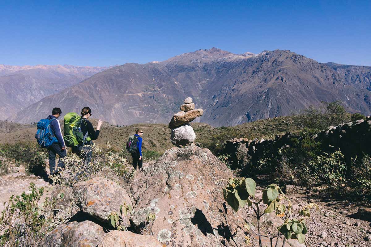 Three trekkers walking with backpacks past rocks into Colca Canyon.