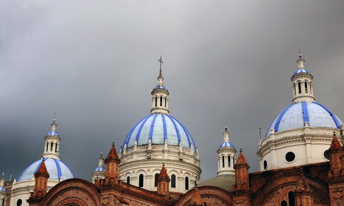 Three pale blue domes on a church in Cuenco, Ecuador against a dark grey sky.