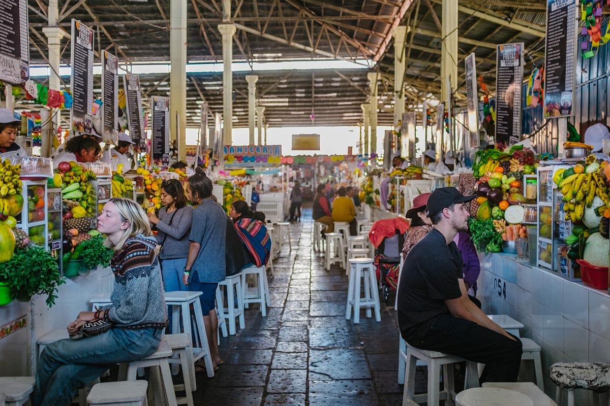 Two vegetarians in Peru sit on stools in front of juice vendors in the San Pedro market in Cusco.