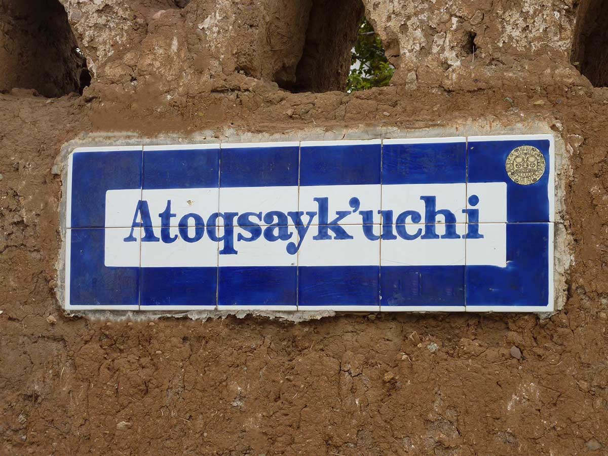 White street sign with blue border that says "Atoqsayk'uchi" on brown wall in Cusco.