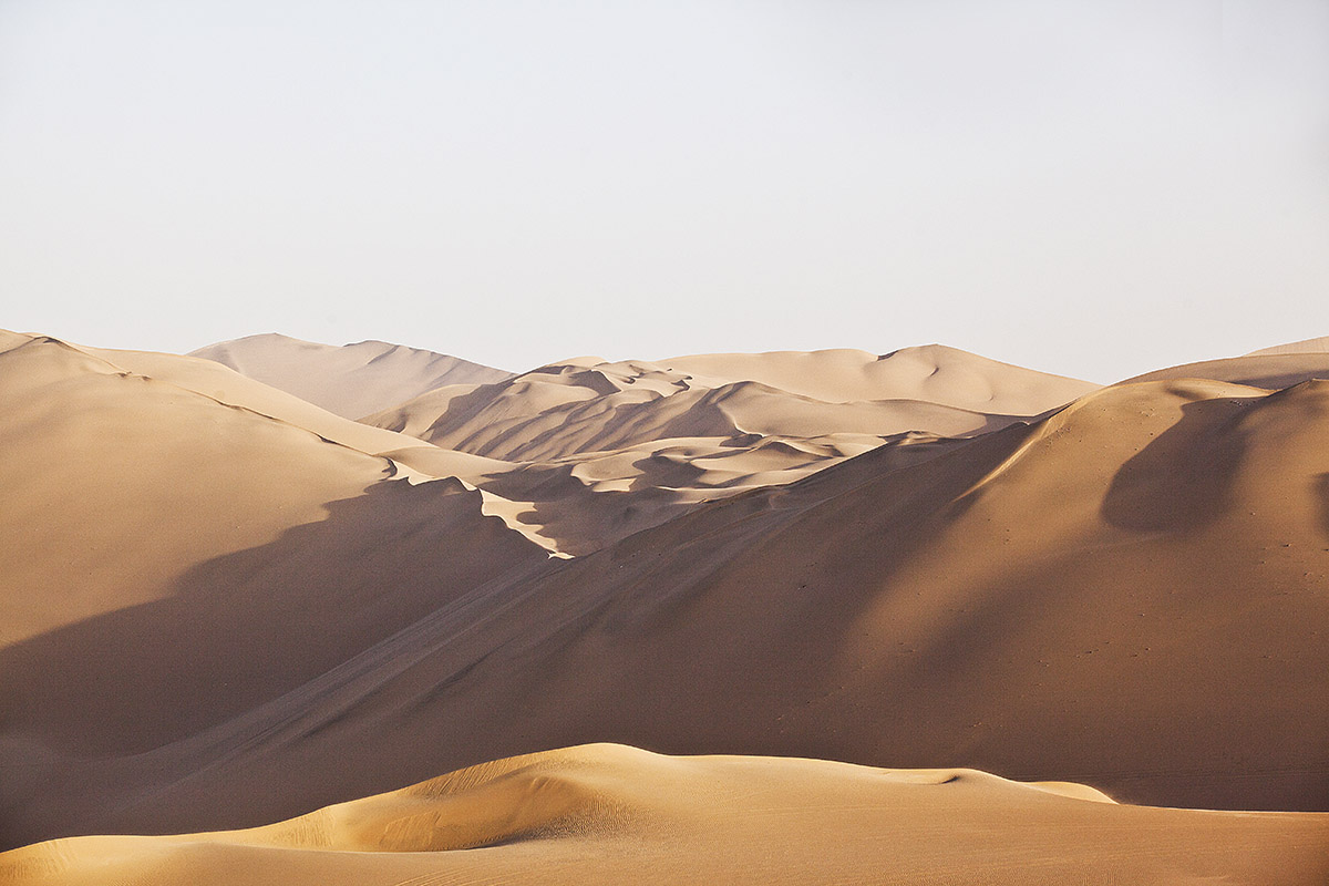 Hilly orange sand dunes under a white sky in the Peruvian Coastal Desert in South America.