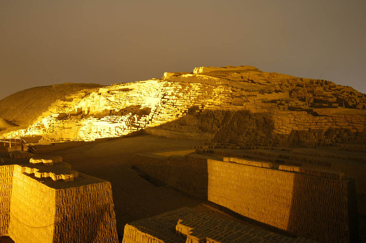 Huaca Pucllana ruins illuminated at night. The lights cast large shadows across the adobe bricks.