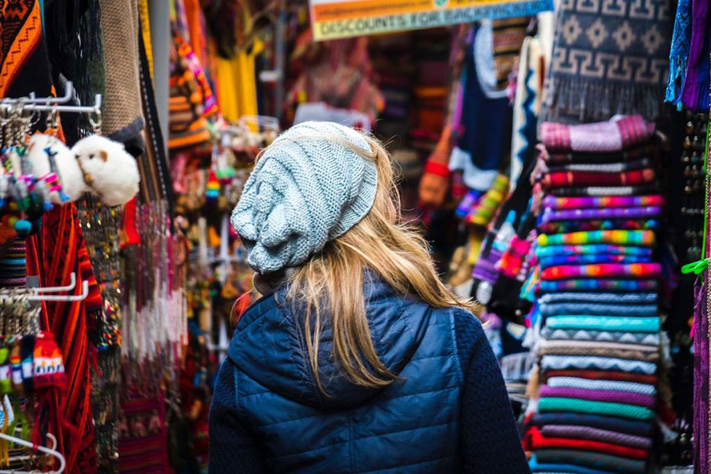 A woman wearing a blue hat is walking through a colorful Inka Market. She is facing away from the camera, showing only her back