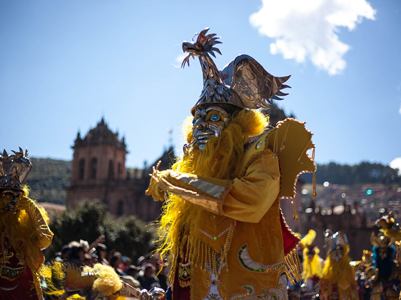 A masked performer at Inti Raymi wearing a costume and a hat with a statue of a creature on it.