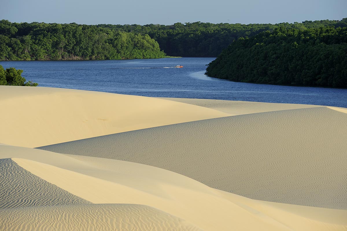 Beige sand dunes back up to a blue river with lush green trees on the other side.