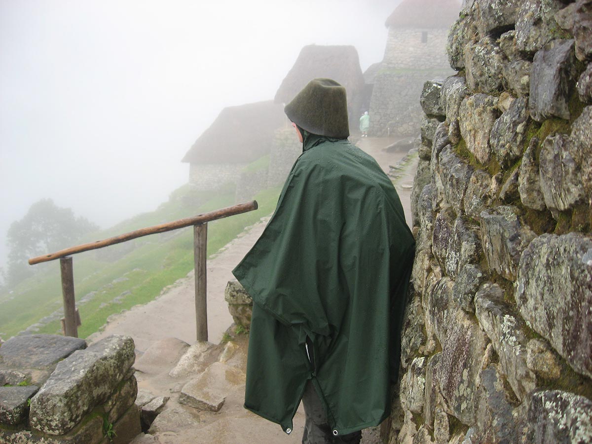A visitor to Machu Picchu in dark green poncho and bucket hat standing next to an Incan wall.