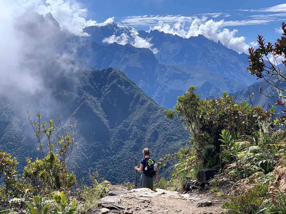 Hiker walking down a path in Machu Picchu with mountains and clouds towering in the background.