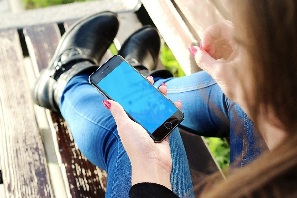A hand holds a black cell phone while sitting. The person is wearing jeans and black boots.