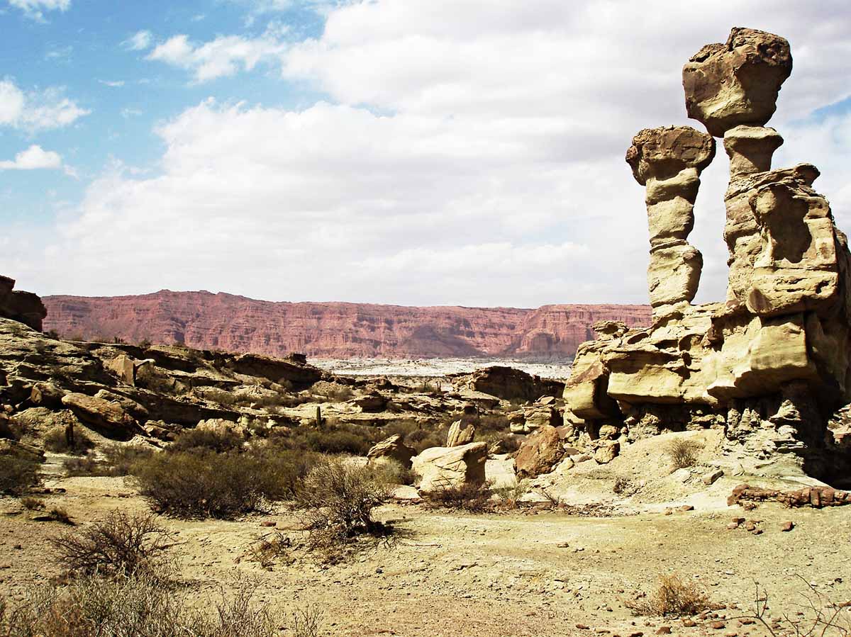 Two tall rock formations surrounded by barren, rocky terrain with red hills in the distance.