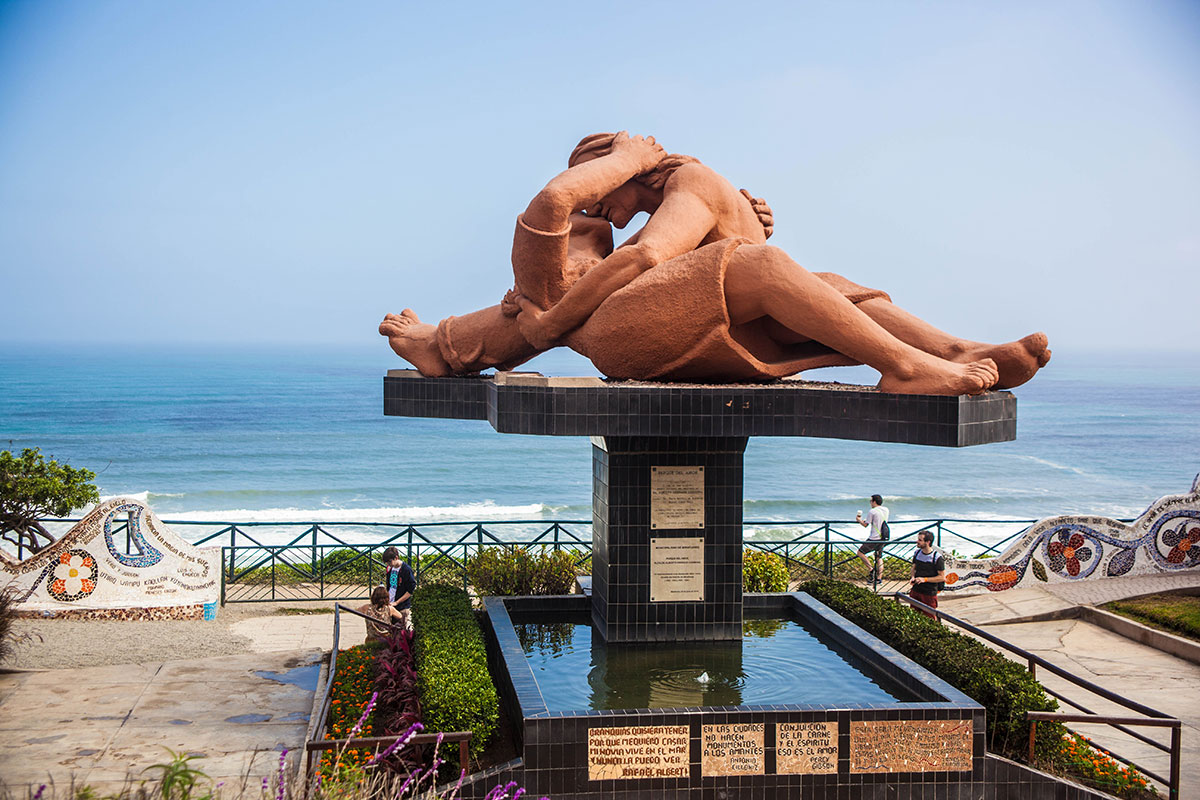 A large light orange statue sculpted to show a man and woman sitting down and embarrassing each other for a kiss. Behind the sculpture is the ocean.