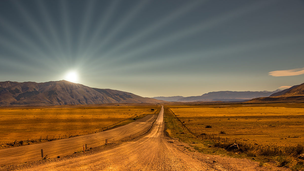 Brown, empty terrain with a dirt road running through. Mountains with a sun setting in the distance.