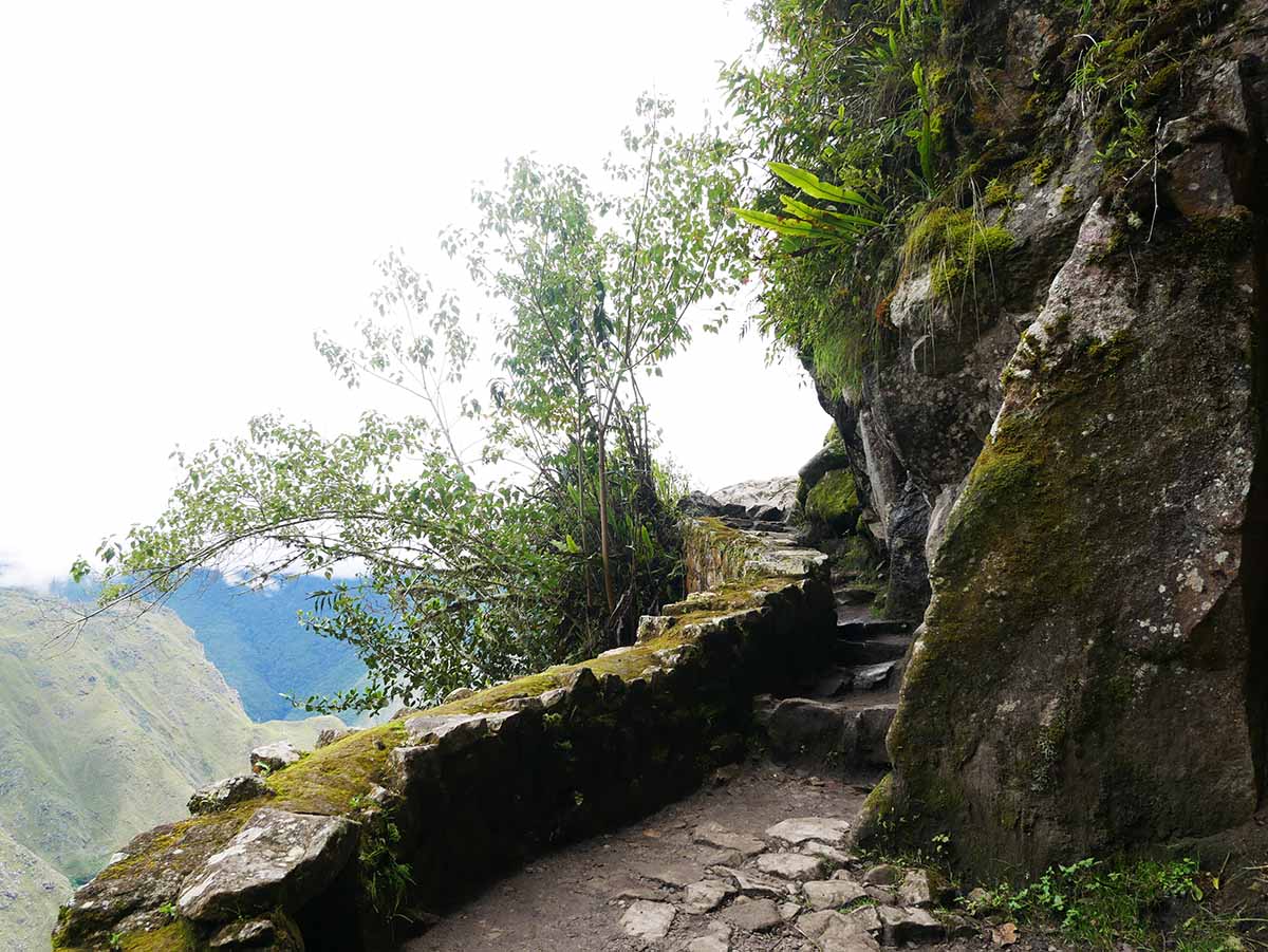A narrow, winding stone path leads to the Inca Bridge along the side of a mountain.