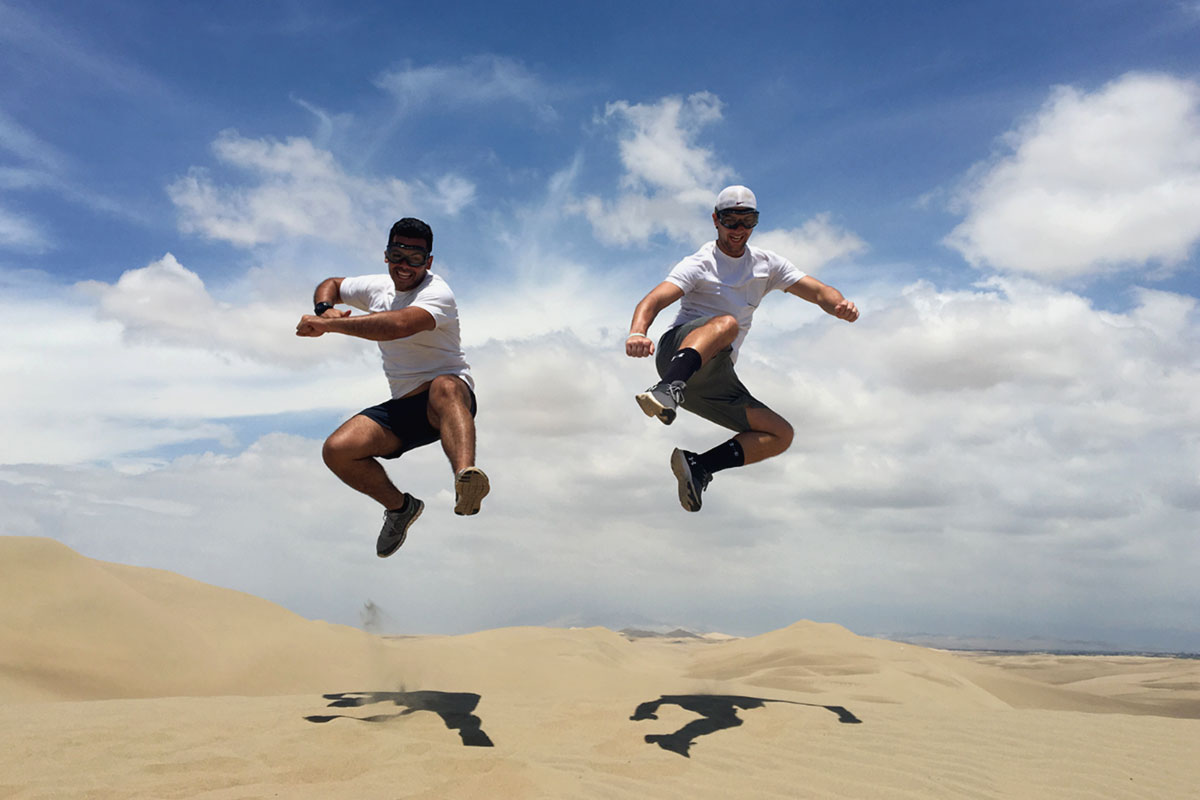 Two men happily jumping in the desert of Peru. It is a clear day and the sand below them is a light brown. They are both wearing shorts and white t-shirts.