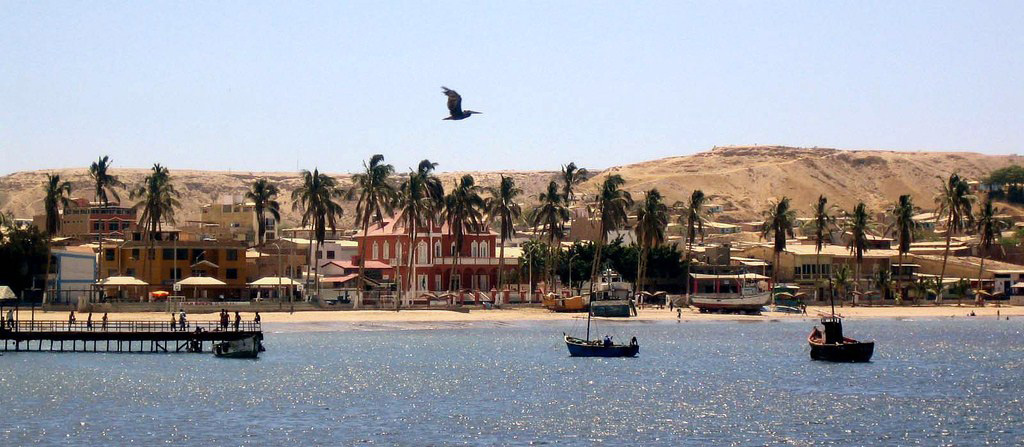 Boats and docks in the ocean with buildings and palm trees on the land. Sandy desert terrain behind.