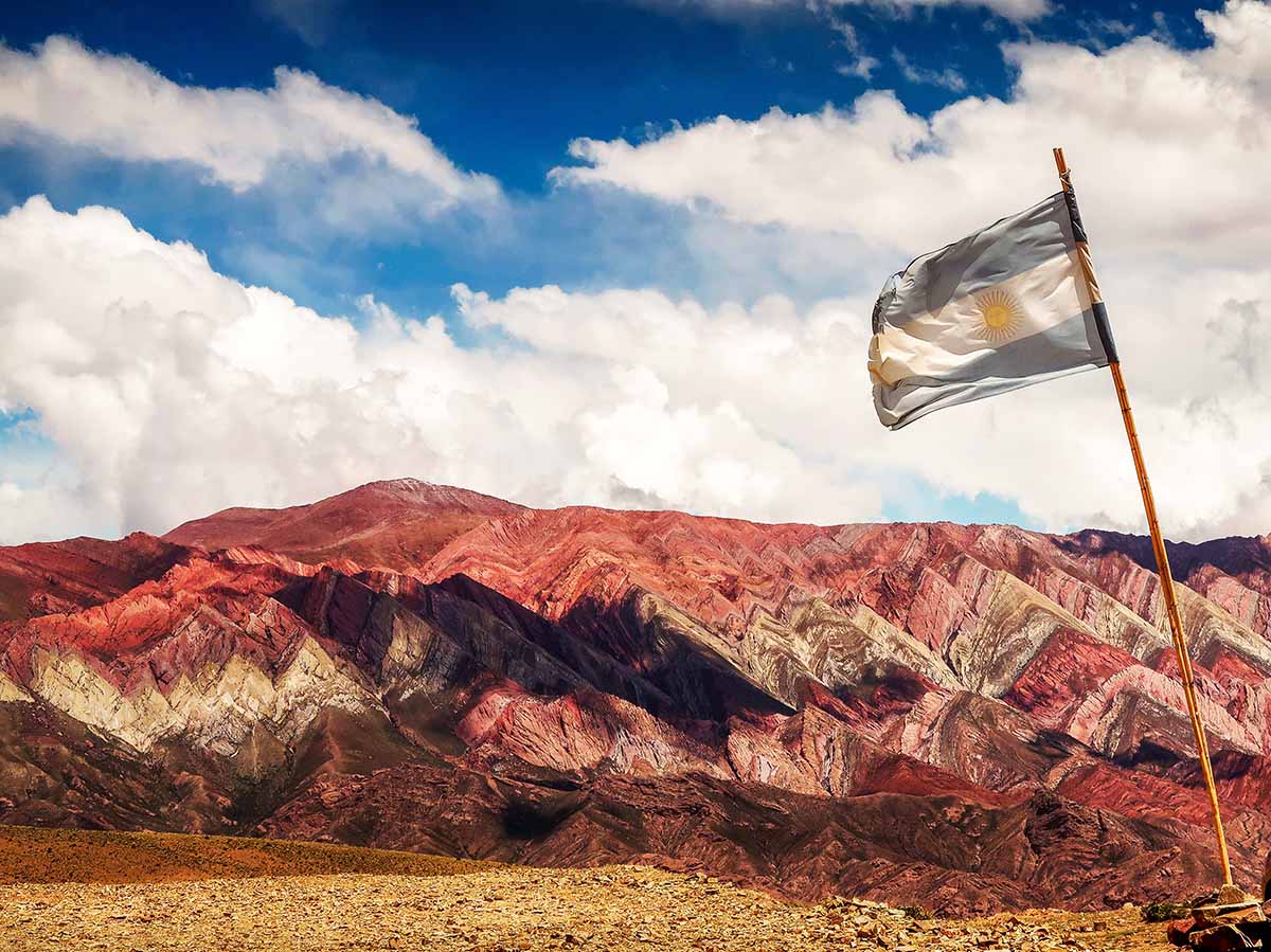 Rocky mountains with hues of red and brown under cloudy skies. An Argentinean flag flies above.