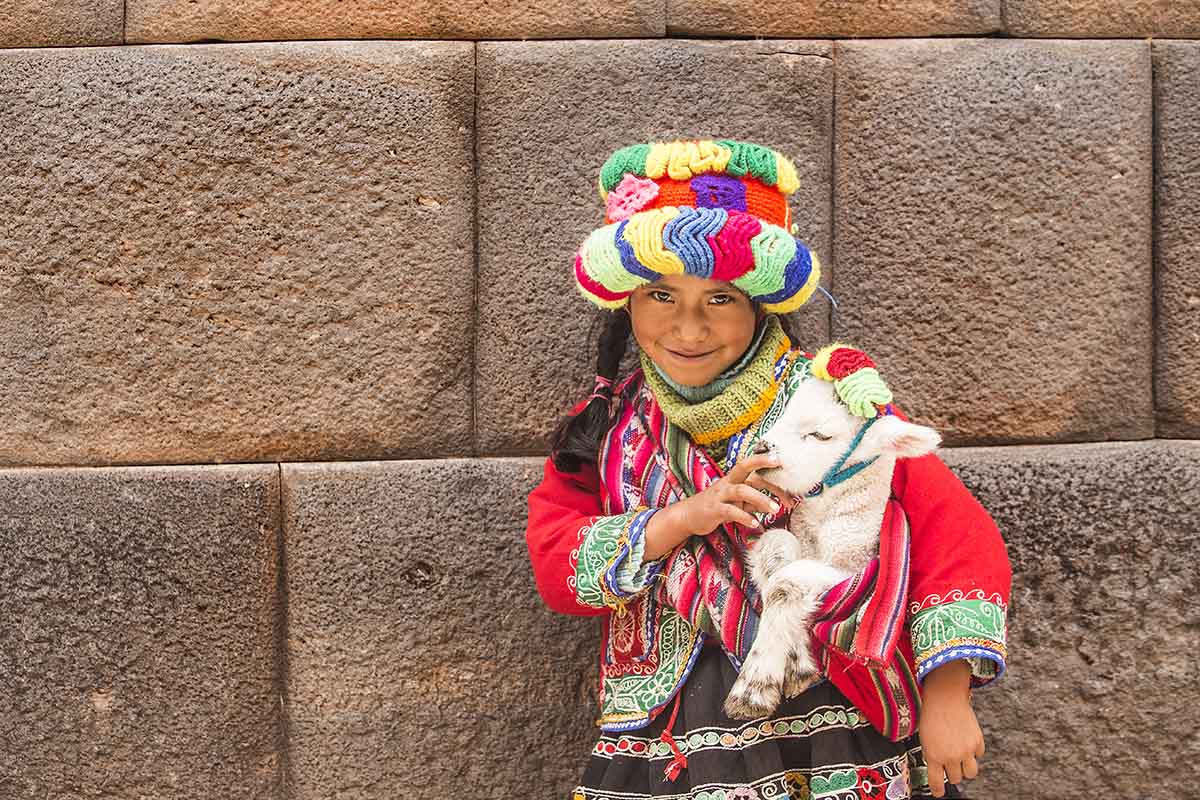 Young Quechua girl in multicolored traditional dress holding a baby lamb.