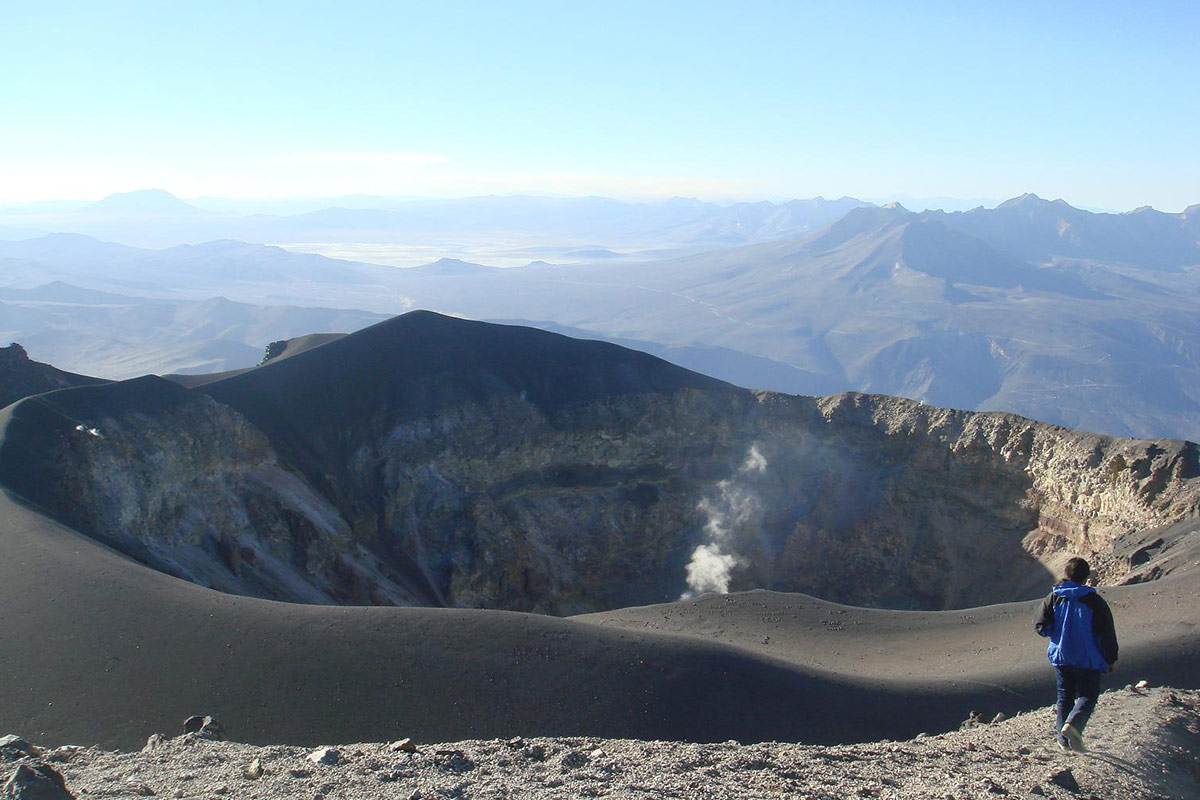 The circular crater summit of Misti Volcano with some smoke coming out and a trekker looking over.