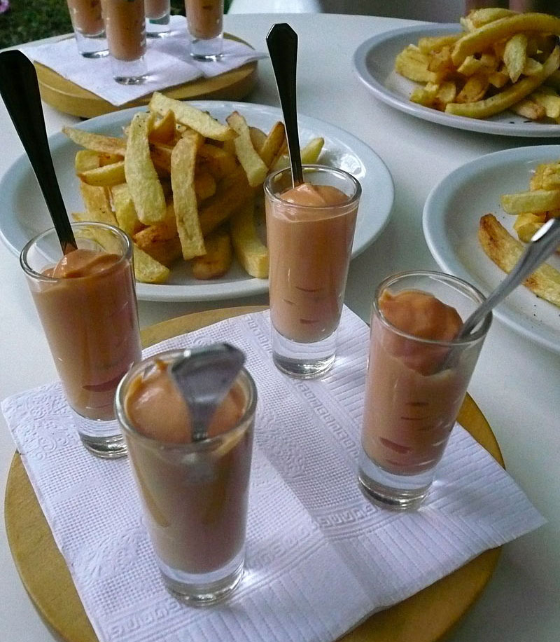 Four glass cups with salsa golf and silver spoons. Plates with fries and more sauce cups behind.