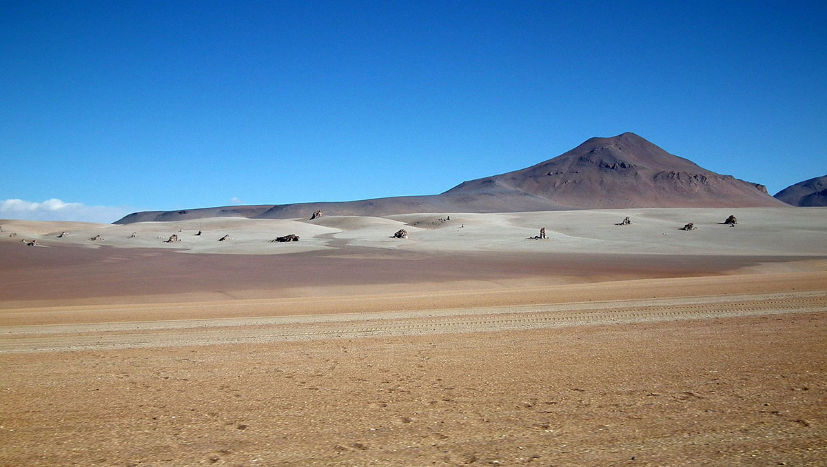 A barren, sandy desert with a mountain in the horizon under blue skies.