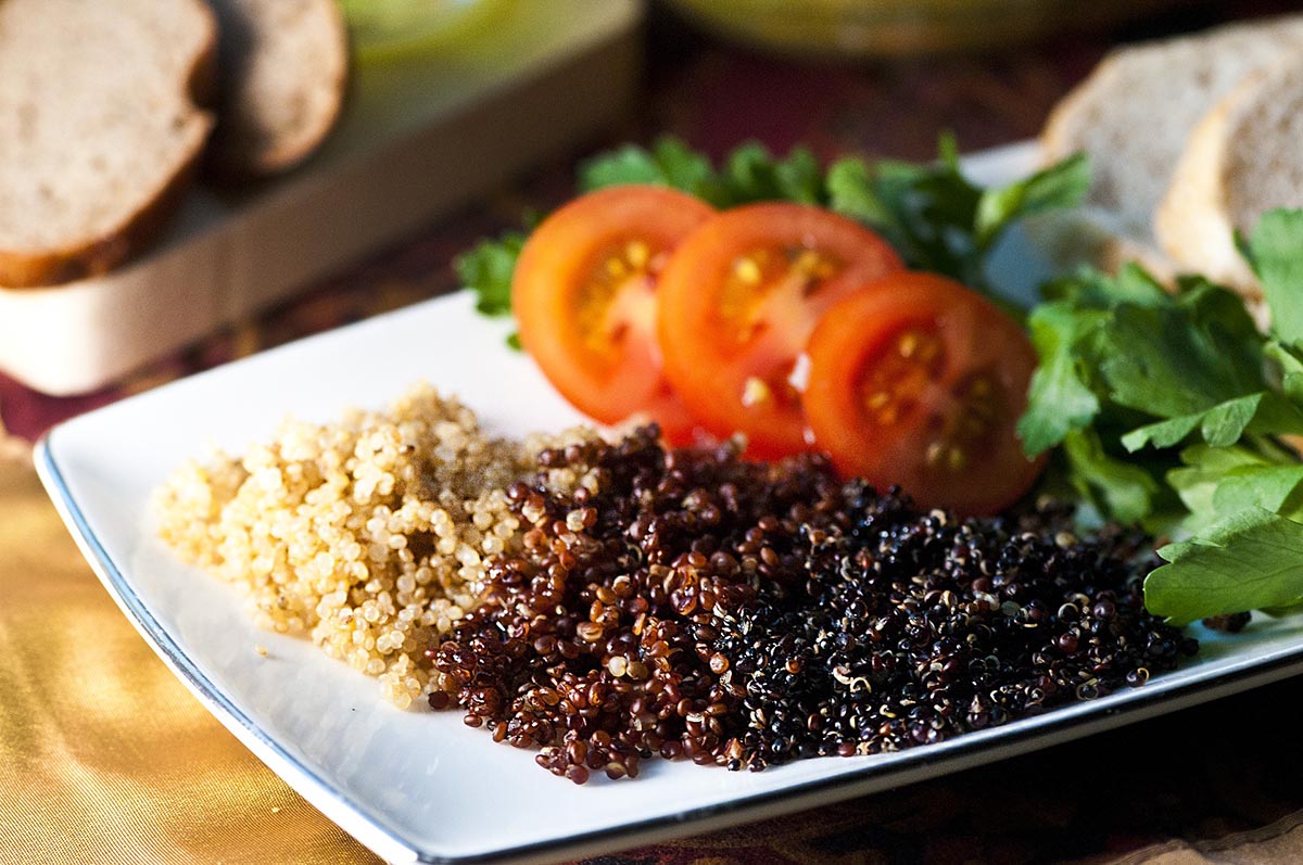 Cooked red, white and black quinoa on a white plate with tomatoes and greens..