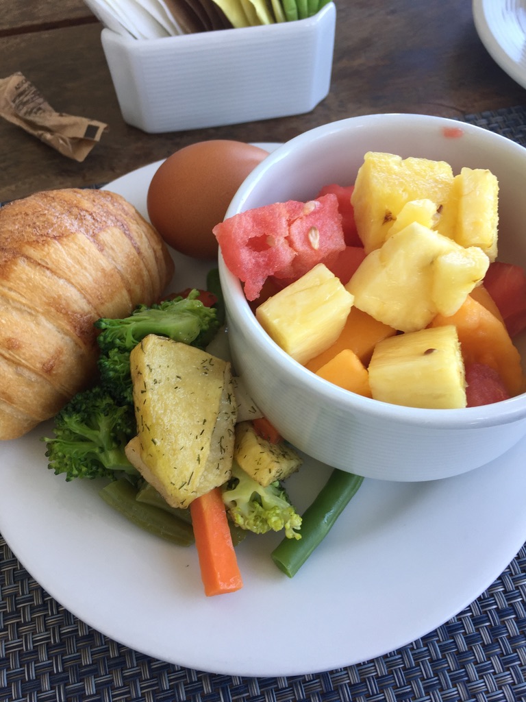 A vegetarian breakfast of fruits, vegetables, a pastry, and a hard boiled egg on a white plate.